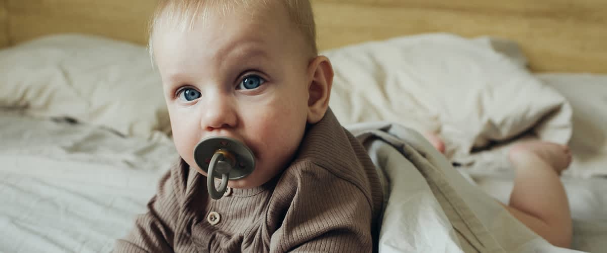 Otrok v rjavem puloverju leži na postelji z dudo v ustih