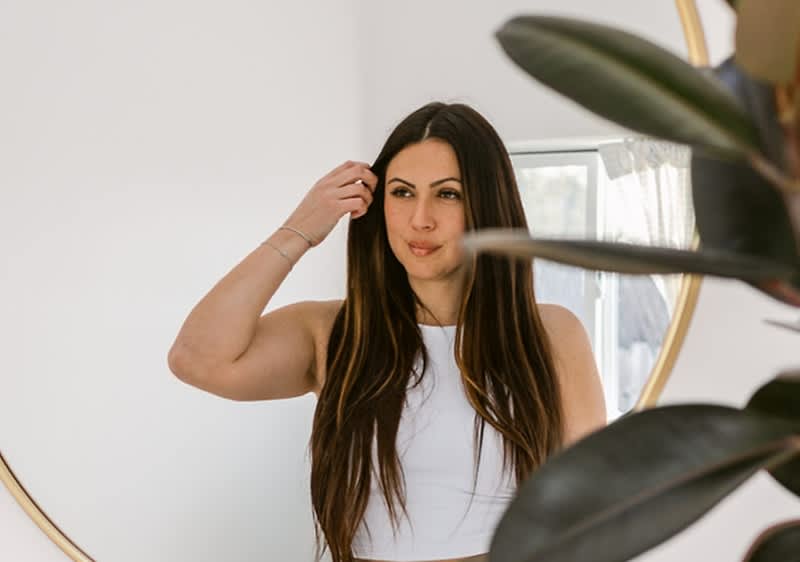 Ženska z dolgimi rjavimi lasmi pred ogledalom, zamegljen obraz, rastlina v ospredju