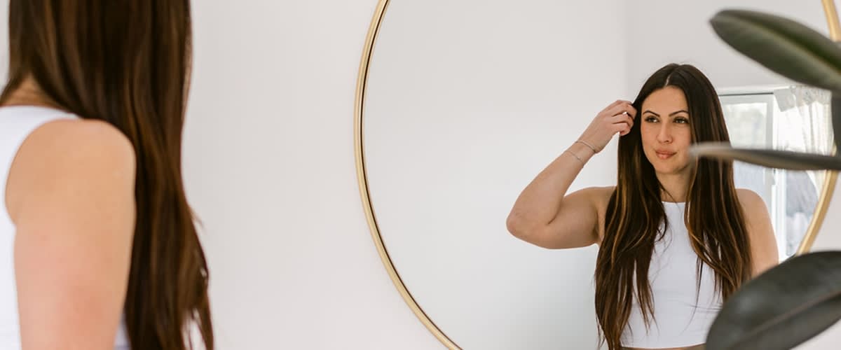 Ženska z dolgimi rjavimi lasmi pred ogledalom, zamegljen obraz, rastlina v ospredju