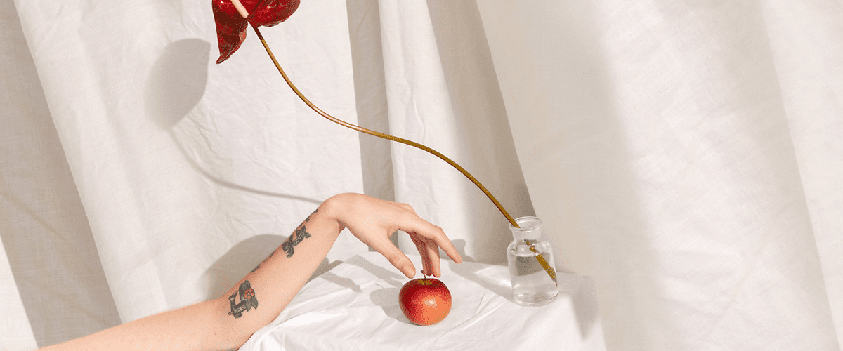 Alt-Text Roka s tetovažo drži jabolko ob vazi z rdečim cvetom na belem ozadju