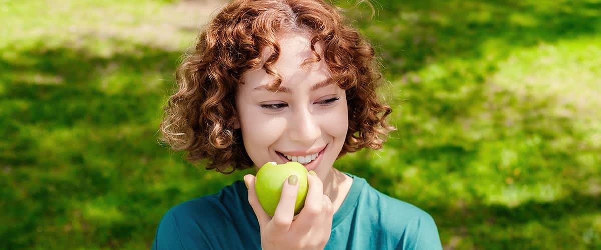 Alt-Text: Oseba z rdečimi kodri drži zeleno jabolko na travniku