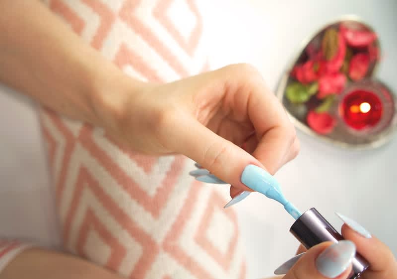 Alt-tekst: Oseba nanaša svetlo moder lak na nohte, v ozadju srčna posoda z dišavnim cvetjem.