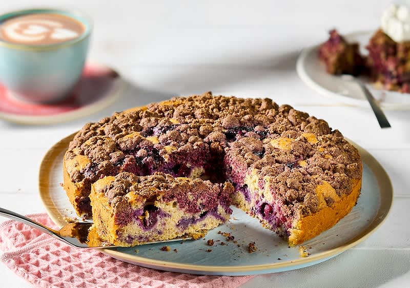 Alt-tekst: Borovničeva torta s hrustljavim posipom na krožniku, ob skodelici kave.