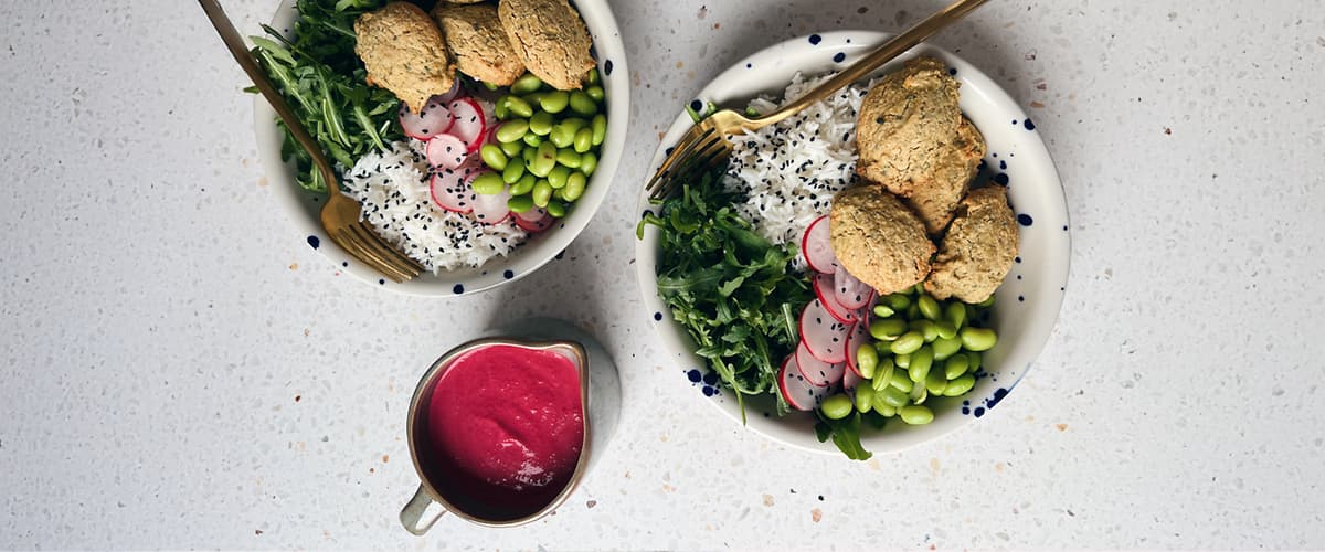 Alt-tekst: Dve skledi z rižem, falaflom, redkvicami, edamamom in rukolo ter skodelica rdeče omake.