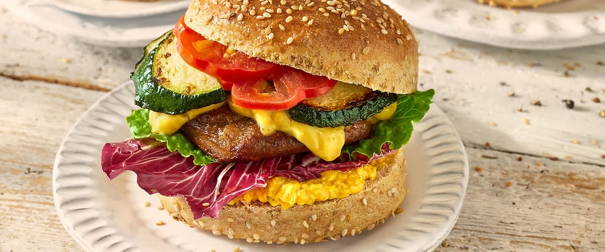 Alt-tekst: Hamburger z zelenjavo, sirom in solato na beli krožniku.