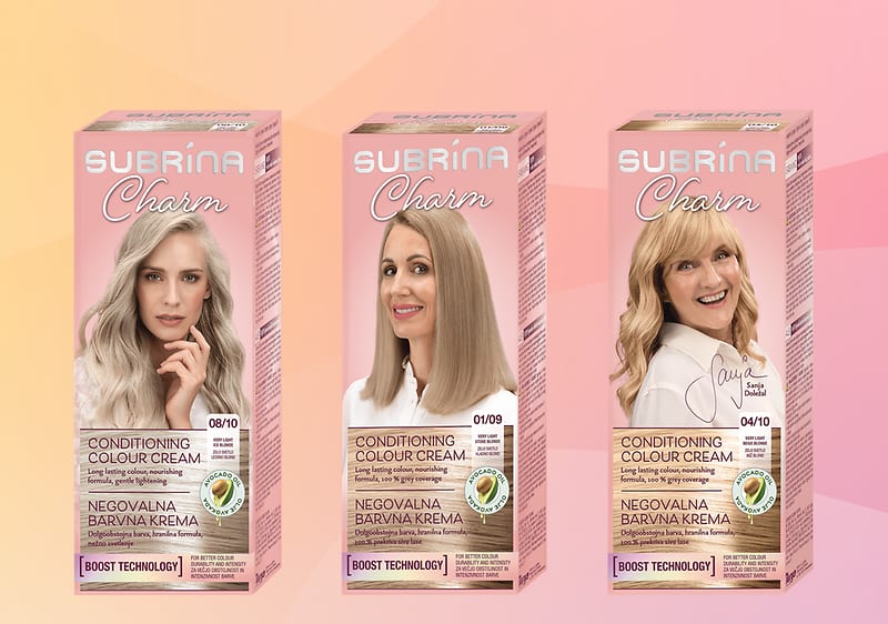 Ženska z blond lasmi nasmejana ob škatlicah barve za lase Subrina Charm, embalaže prikazane spredaj