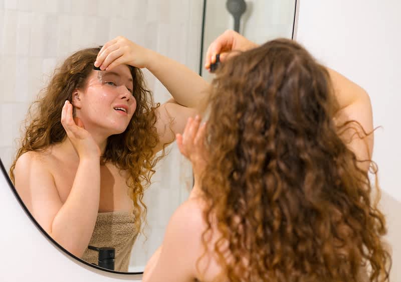 Ženska nanaša serum na obraz pred kopalniškim ogledalom, zavit v brisačo