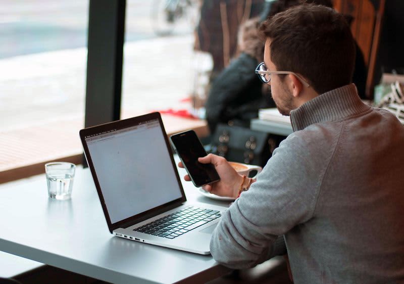 Mann sitzt vor seinem Laptop und wählt eine Nummer auf seinem Smartphone.