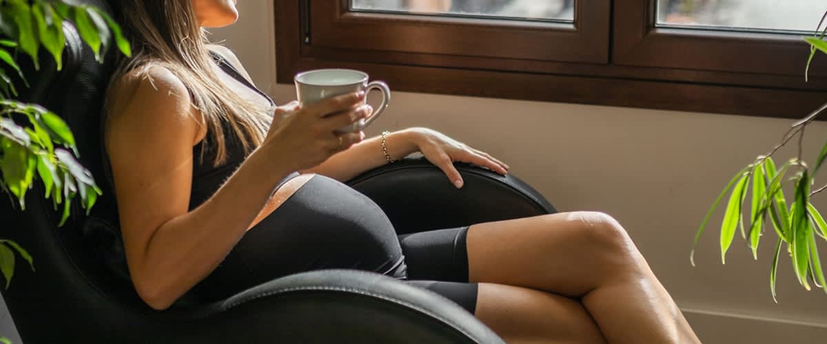 Schwangere Frau sitzt mit einer Tasse Tee in der Hand am Fenster