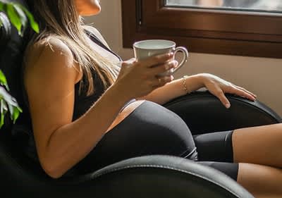 Schwangere Frau sitzt mit einer Tasse Tee in der Hand am Fenster