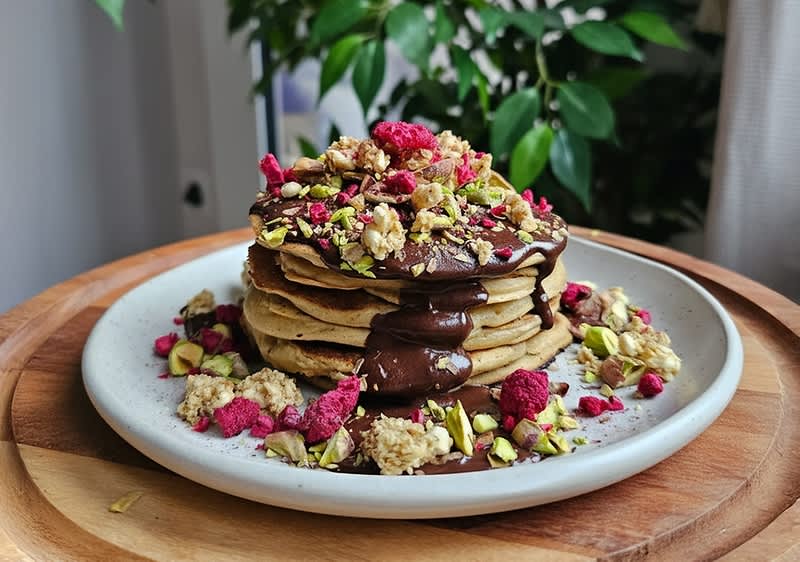Ein Stapel Pfannkuchen mit Schokoladensauce, garniert mit Pistazien, gefriergetrockneten Himbeeren und Müsli auf einem weißen Teller.