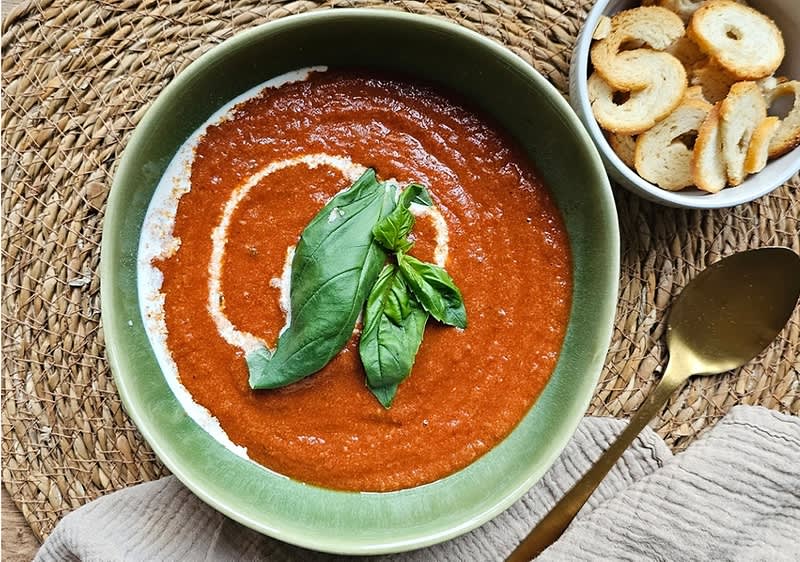 Tomatensuppe in einer grünen Schüssel, garniert mit Basilikumblättern und einem Sahnestrich, daneben eine Schale mit Brotchips.