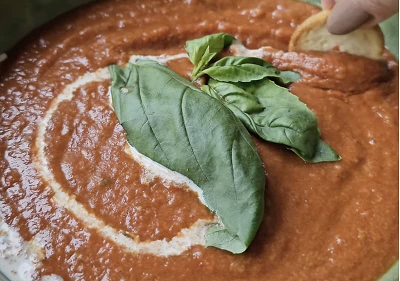 Tomatensuppe mit Sahne und Basilikumblättern, eine Hand taucht ein Stück Brot hinein